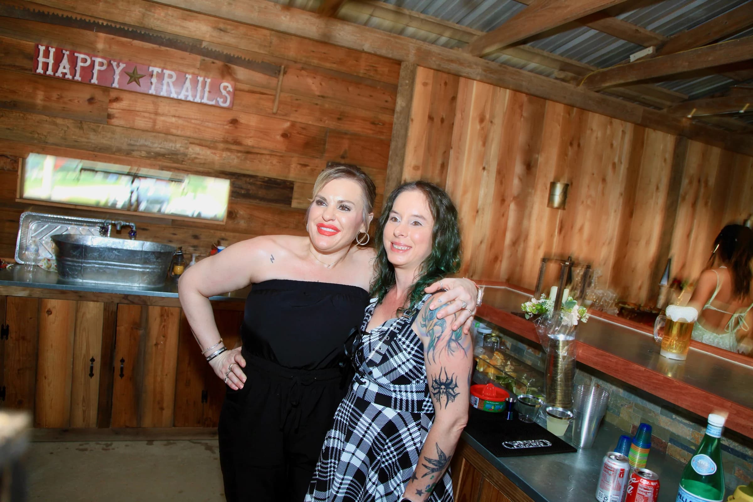 Two smiling women standing beside the wooden bar beneath the Happy Trails sign
