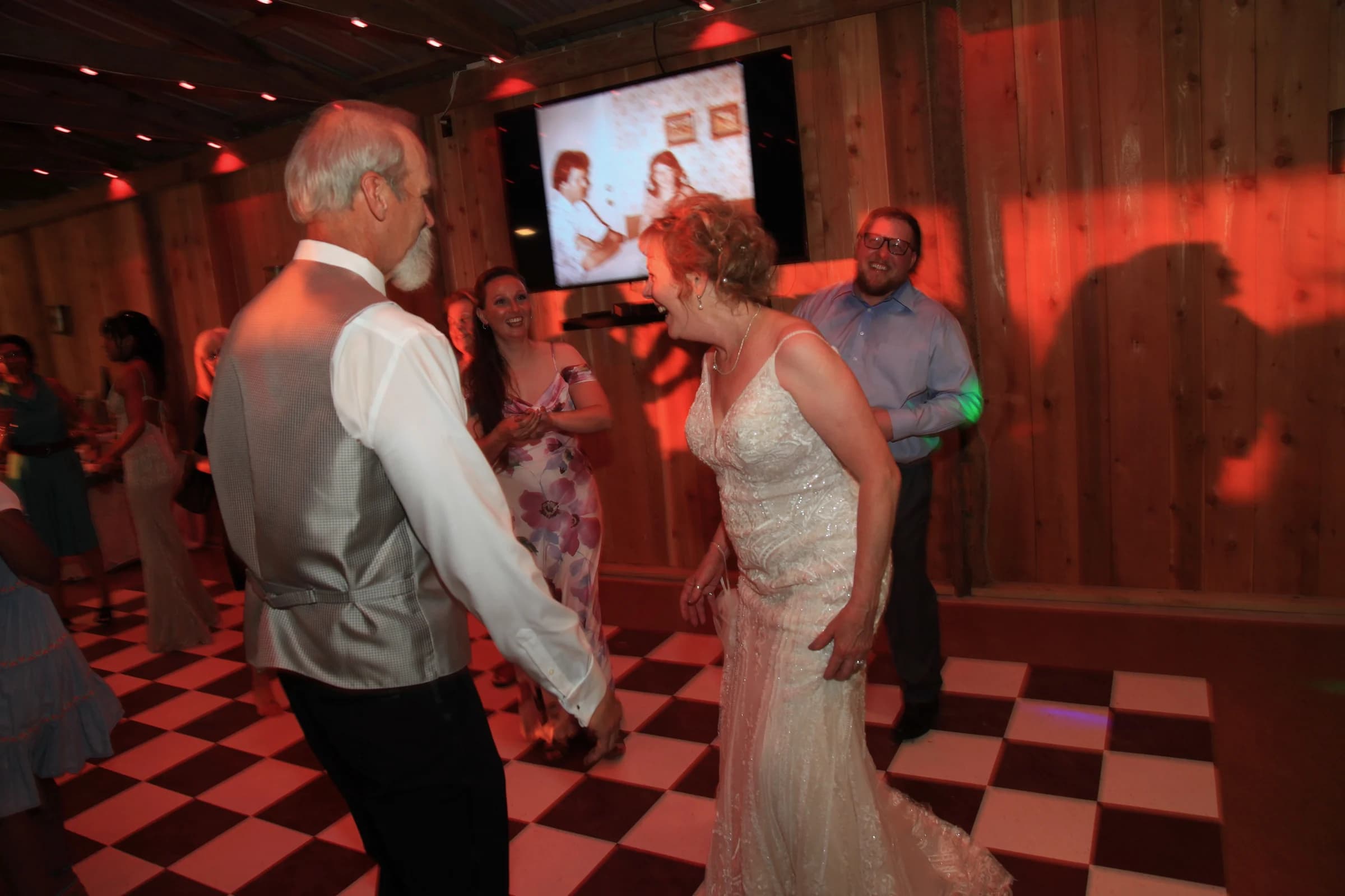 A bride and a man in a vest dancing on the checkered floor, with wedding guests nearby